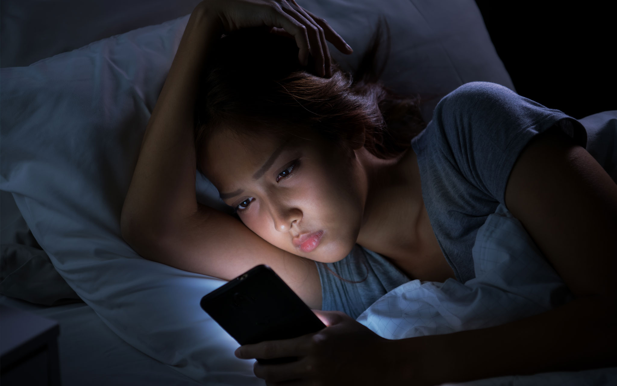 A girl lying on her side in bed in the dark as she views her phone. The light from her phone is illuminating her face.