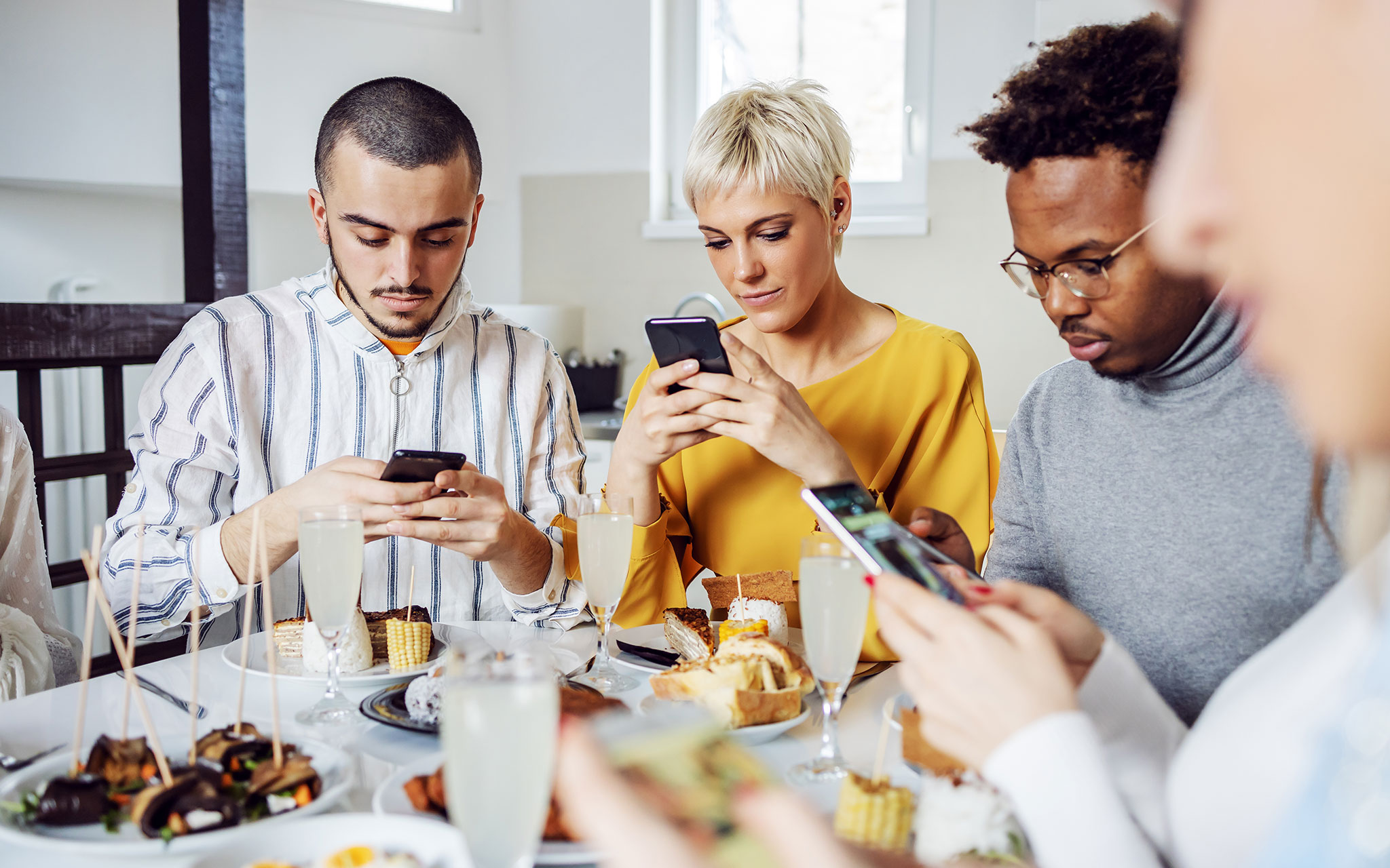 Group of multicultural friends sitting at dinning table for lunch and using smart phones.